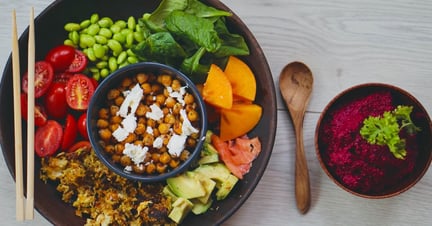A spread of plant-based food: a large bowl containing a smaller bowl full of chickpeas, surrounded by avocados, tomatoes, edamame beans, spinach and sweet potato. Next to it, a smaller bowl filled with bright purple beetroot dip.
